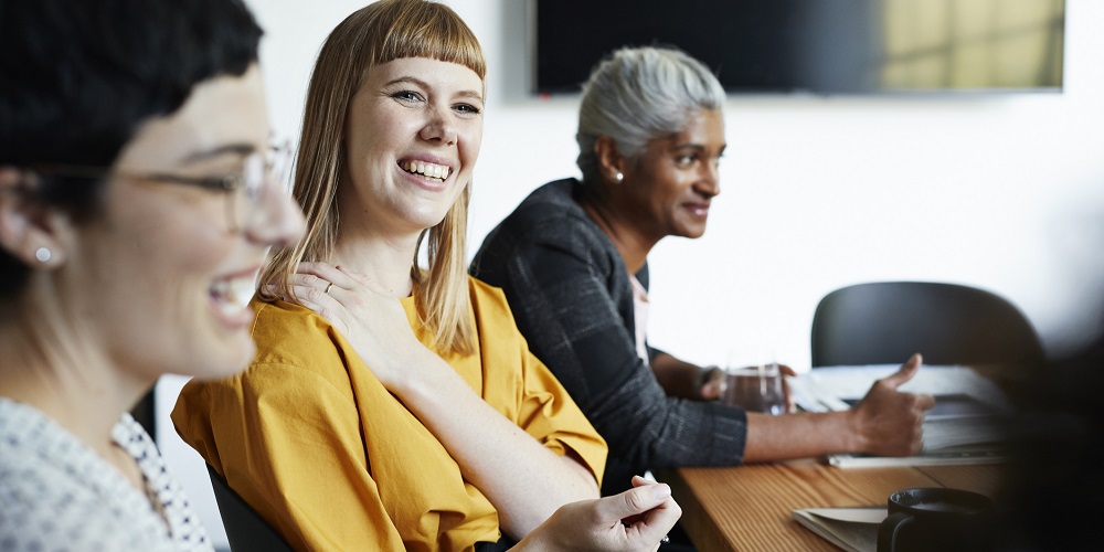 A diverse set of female colleagues is listenting to a presentation at the office together. They are laughing and having a good time.